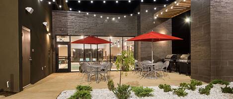 Un patio al aire libre con mesas y sillas, luces colgantes y una parrilla.