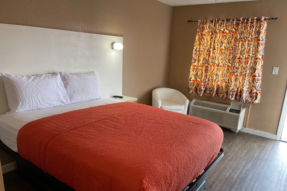 A hotel room with a large bed, white pillows, a red blanket, a white chair, a heater, and a patterned curtain.