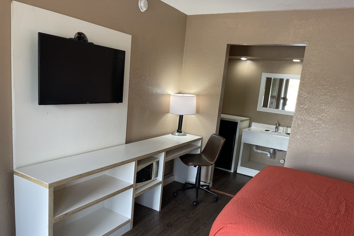 A hotel room with a bed, a desk with a chair, a TV mounted on the wall, a lamp, a mirror, and a sink.