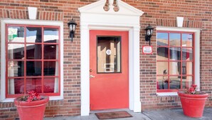 Una puerta roja con detalles blancos, flanqueada por dos ventanas con marcos rojos y un muro de ladrillo.