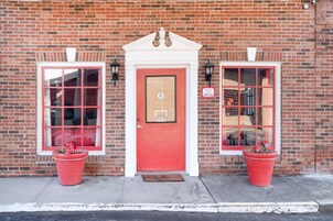 A red door with a white trim, flanked by two windows with red frames, and two potted plants on the sidewalk.