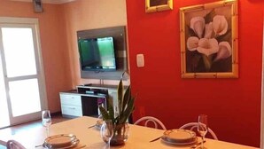 Un coin repas avec une table blanche dressée pour quatre personnes, entourée de chaises blanches. Deux tableaux représentant des motifs floraux, encadrés, ornent le mur, et un téléviseur est fixé au-dessus de la table.