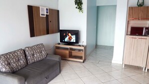 Un salon avec un canapé gris, un téléviseur posé sur un meuble en bois et un petit meuble contenant un four à micro-ondes.