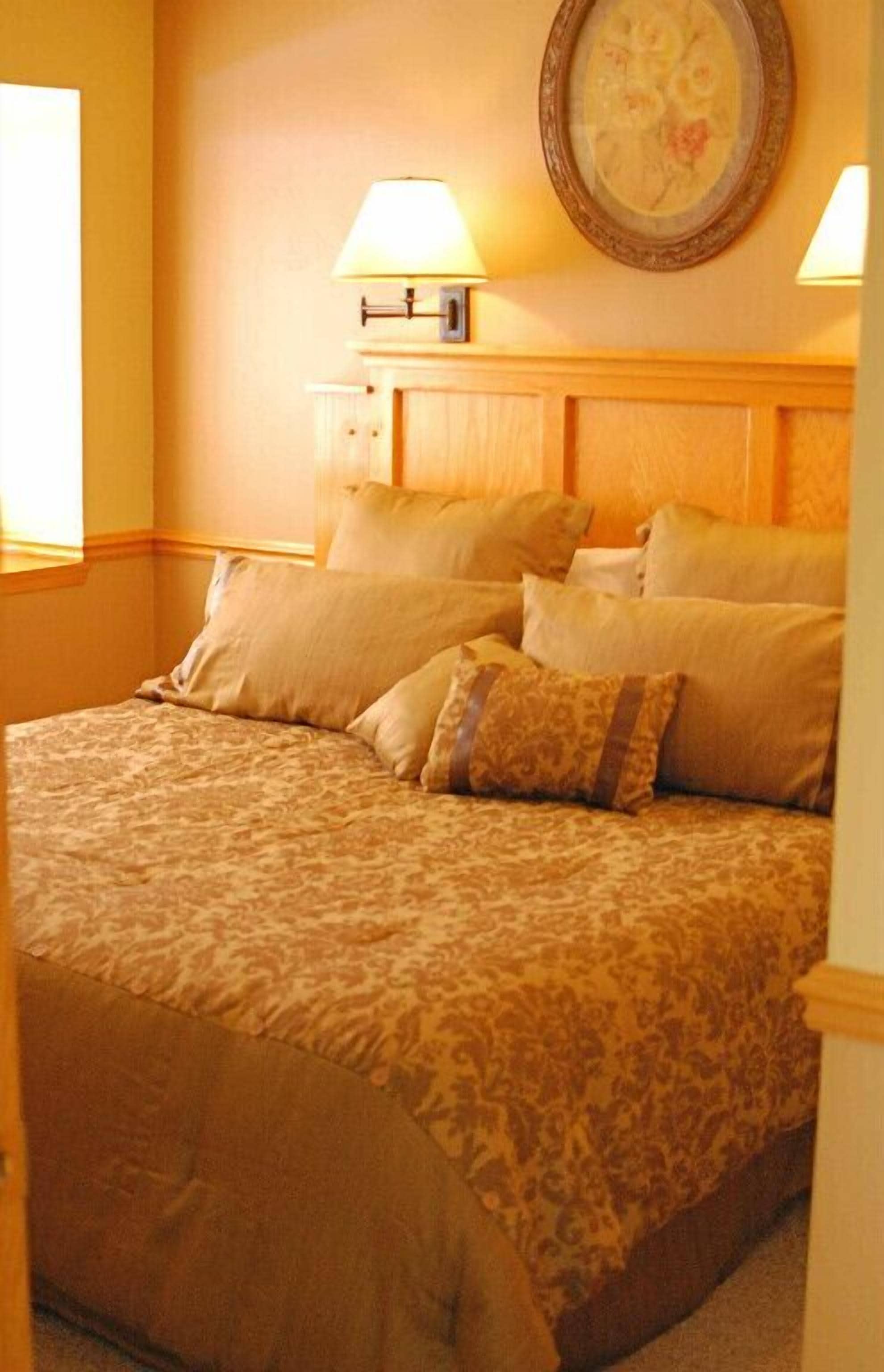 A neatly made bed with a patterned bedspread and several pillows, a bedside lamp, and a framed floral artwork on the wall.