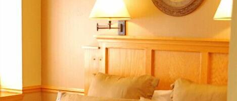 A neatly made bed with a patterned bedspread and several pillows, a bedside lamp, and a framed floral artwork on the wall.