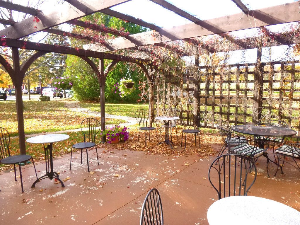 Un patio al aire libre con mesas y sillas, cubierto por una pérgola con plantas trepadoras.