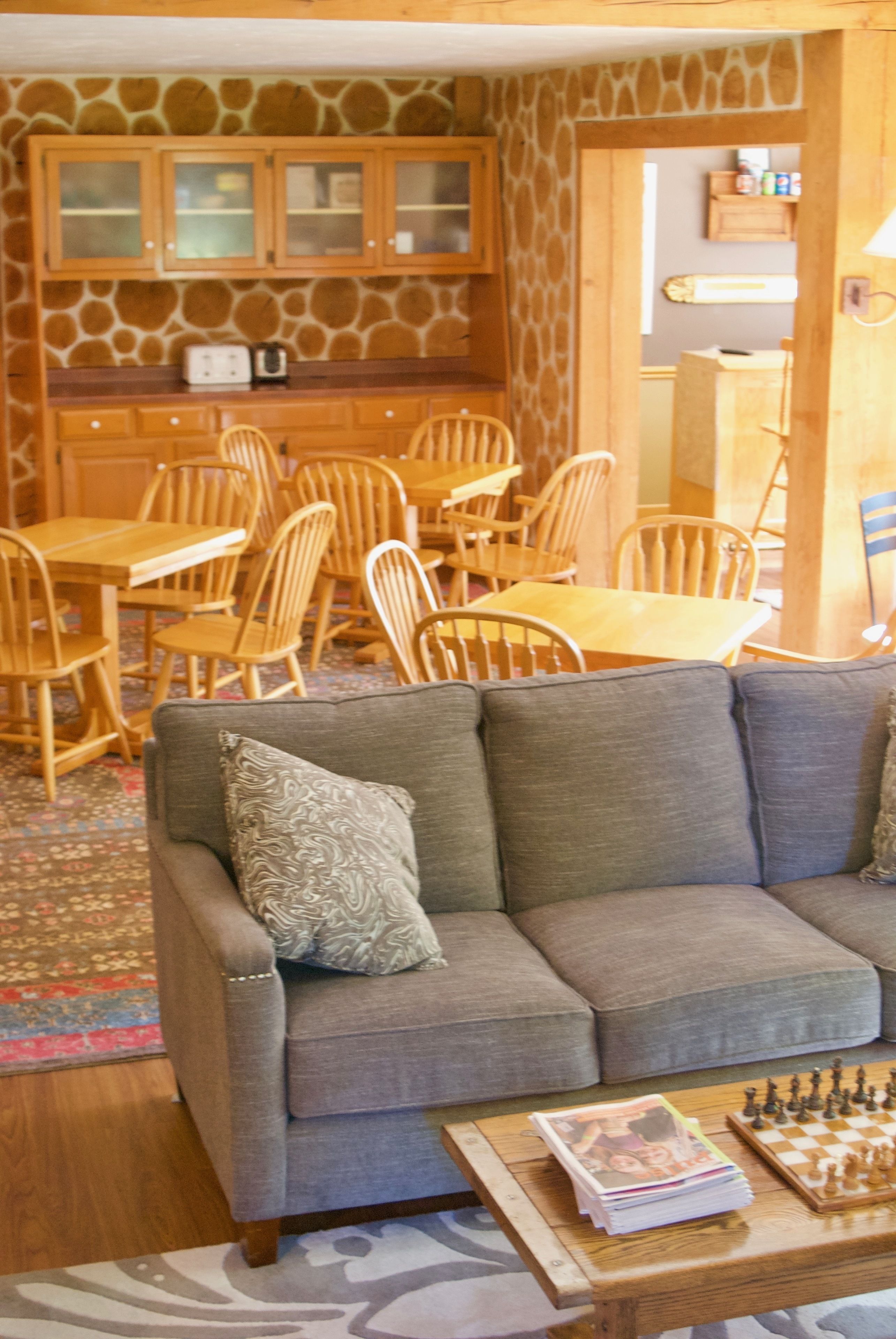 A cozy living room with a gray sofa, wooden chairs, a coffee table with a chess set, and a kitchen area with wooden cabinets and a stone backsplash.