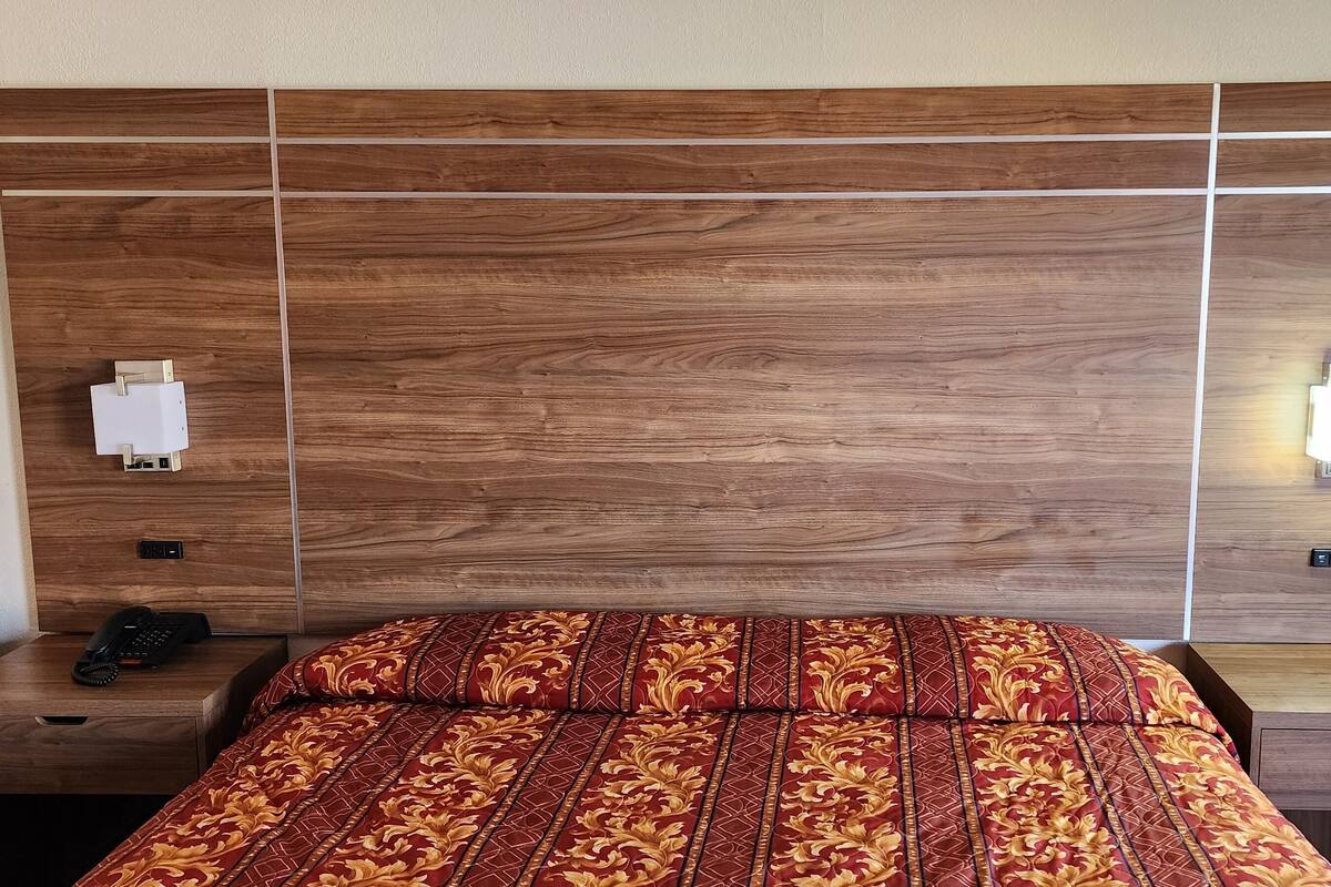A hotel room with a wooden headboard, a bed with patterned bedding, bedside tables with lamps, and a telephone.