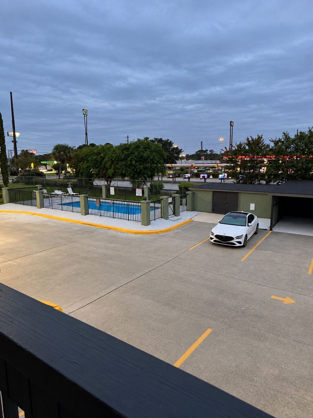 A parking lot with a white car, a swimming pool, and a building in the background.