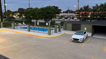 A parking lot with a white car, a swimming pool, and a building in the background.