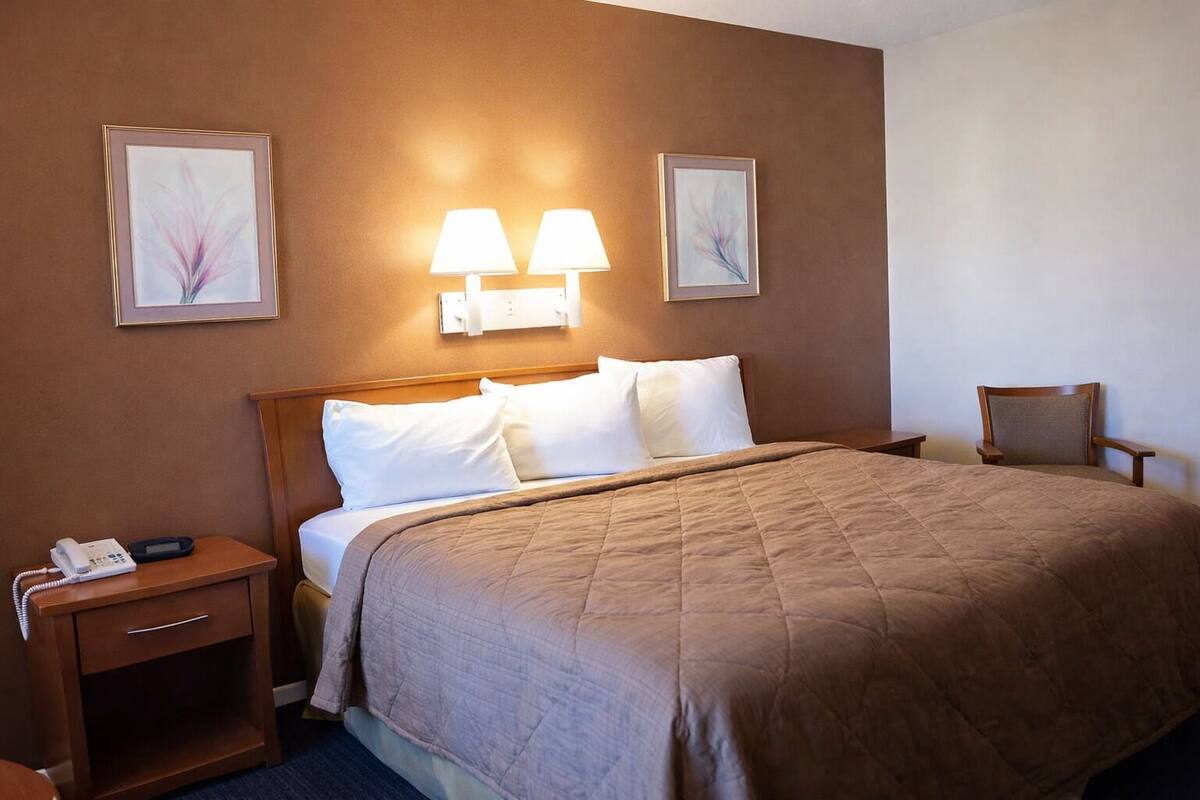 A hotel room with a bed, bedside lamps, a chair, a nightstand with a phone, and two framed pictures on the wall.