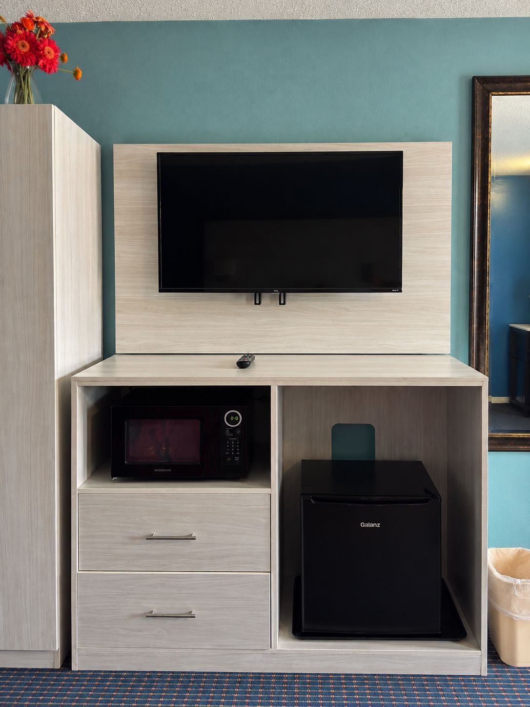 A hotel room with a flat-screen TV mounted on a wooden cabinet, a microwave, a refrigerator, and a trash bin.