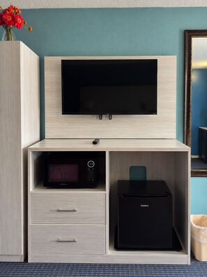 Habitación de hotel con televisor de pantalla plana montado en un mueble de madera, microondas, refrigerador y basurero.