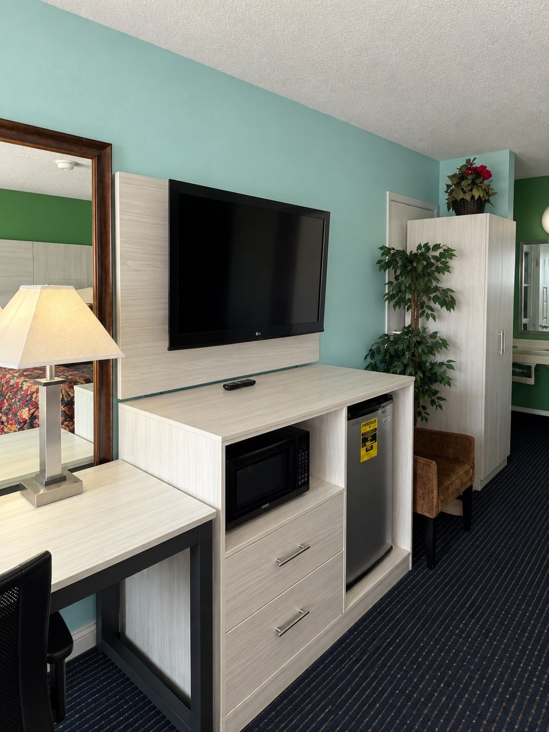 A hotel room with a flat-screen TV mounted on a wall, a microwave, a refrigerator, a desk with a lamp, a chair, and a cabinet with a plant.