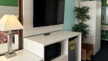A hotel room with a flat-screen TV mounted on a wall, a microwave, a refrigerator, a desk with a lamp, a chair, and a cabinet with a plant.