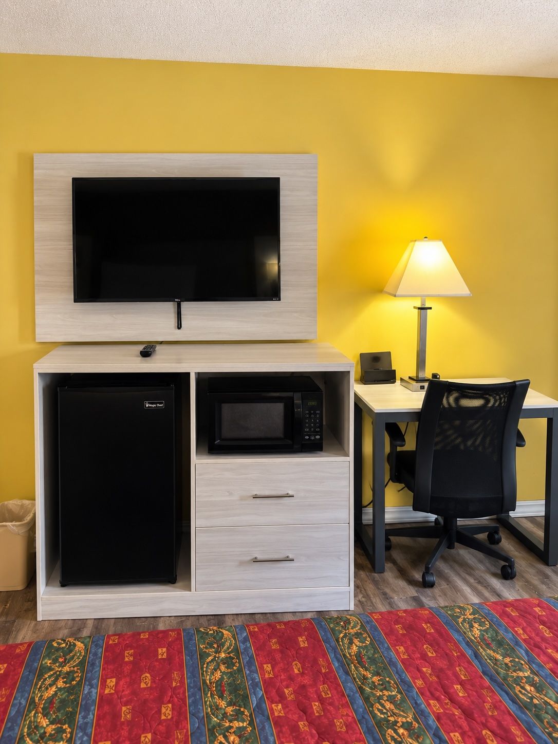 A compact hotel room with a bed, a desk with a lamp, a microwave, and a TV mounted on the wall.
