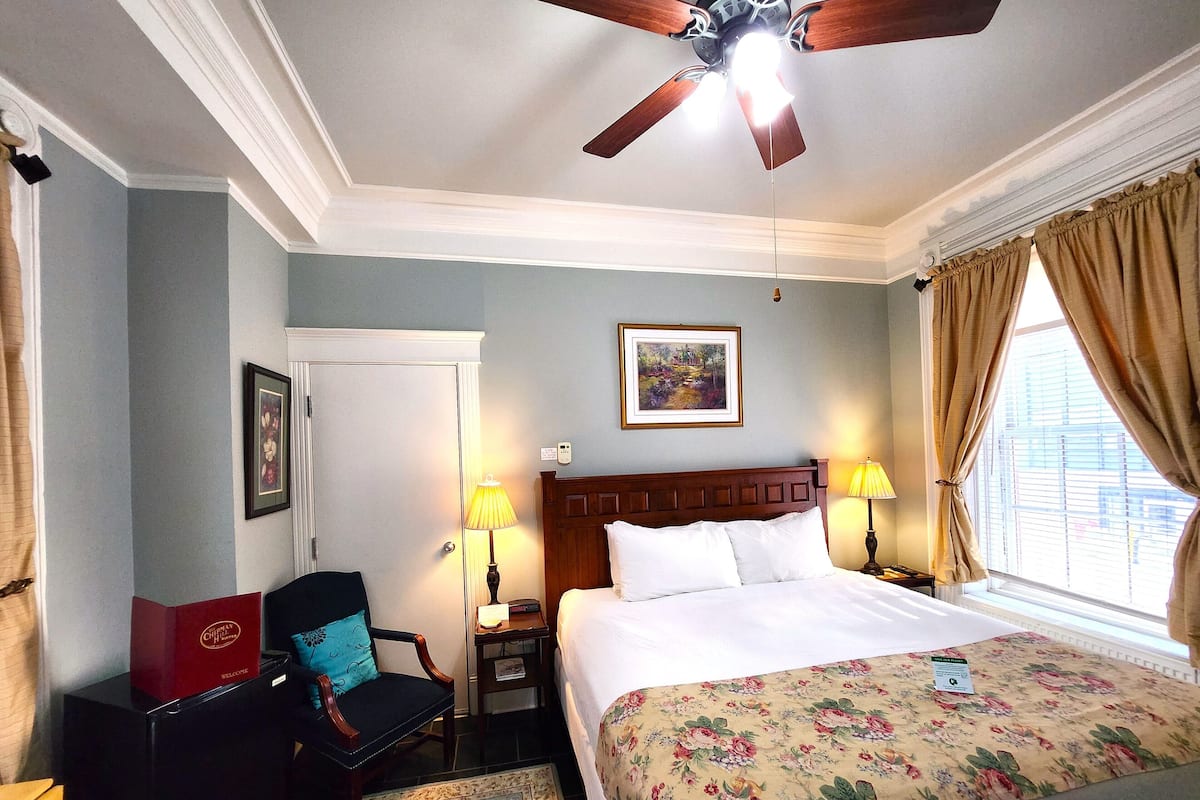A hotel room with a bed, a chair, a desk, a ceiling fan, and a window with curtains.