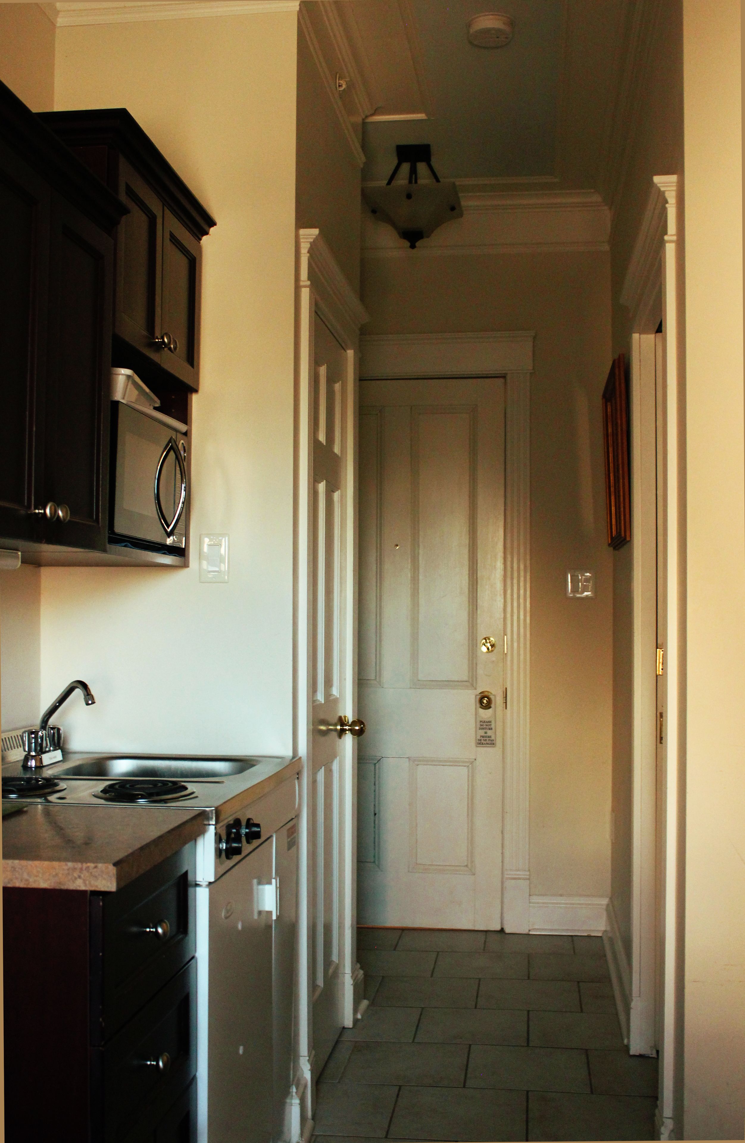 Un couloir étroit, avec un coin cuisine sur la gauche, comprenant une cuisinière, un évier et des placards. Une porte blanche se trouve au bout du couloir.