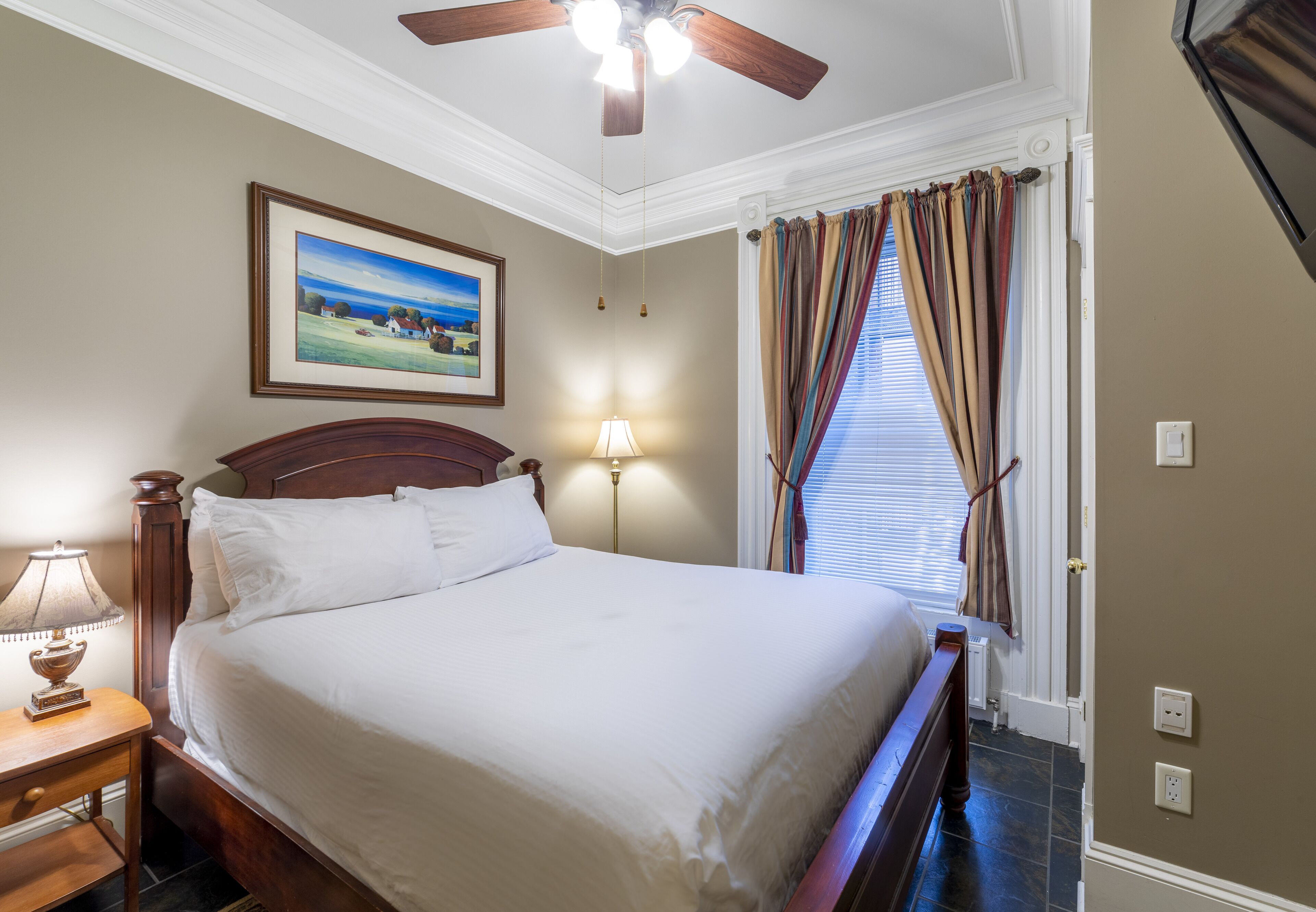 A neatly made bed in a hotel room with a ceiling fan, a framed landscape painting, and a window with curtains.