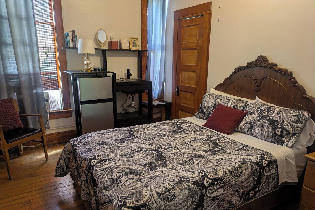 A bedroom with a bed, a desk, a chair, a refrigerator, and a window with curtains.