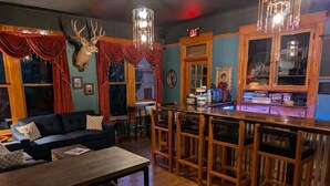 Un coin bar accueillant, avec un comptoir en bois, des tabourets et une tête de cerf accrochée au mur. On y trouve également un canapé et une table avec des bancs, placés au premier plan.