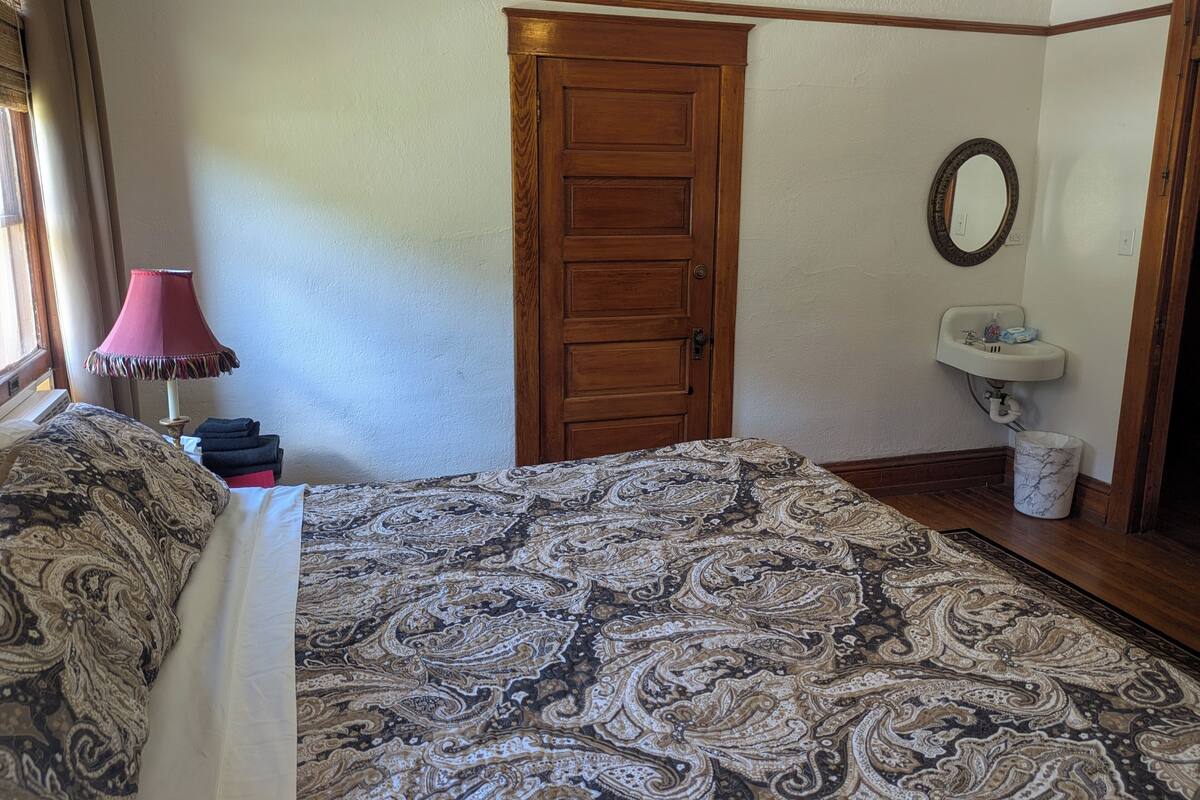 A bedroom with a patterned bedspread, a wooden door, a bedside lamp, a mirror, and a sink.