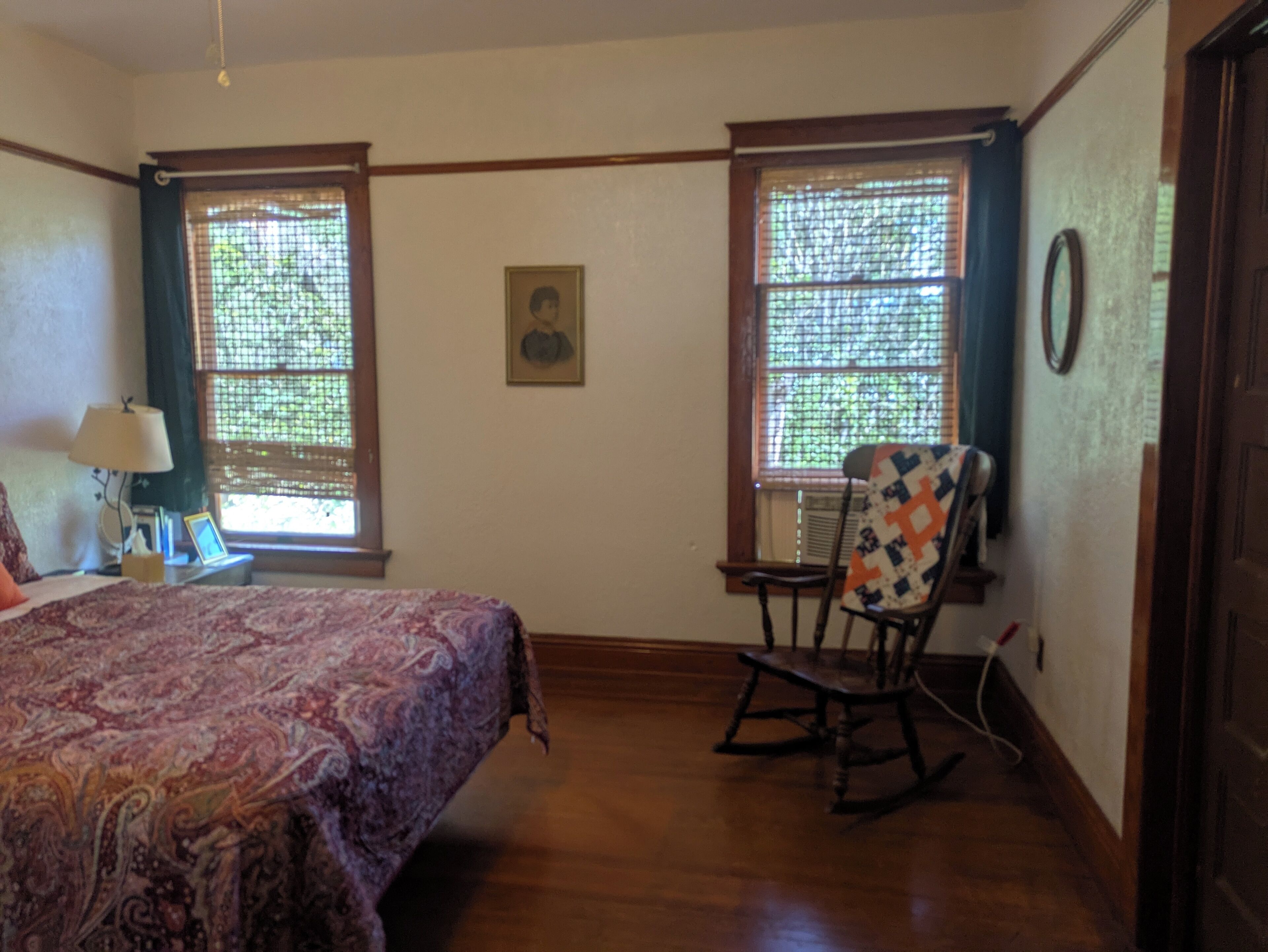 Un dormitorio con una cama, dos ventanas con cortinas, una mecedora, una lámpara y un cuadro en la pared.