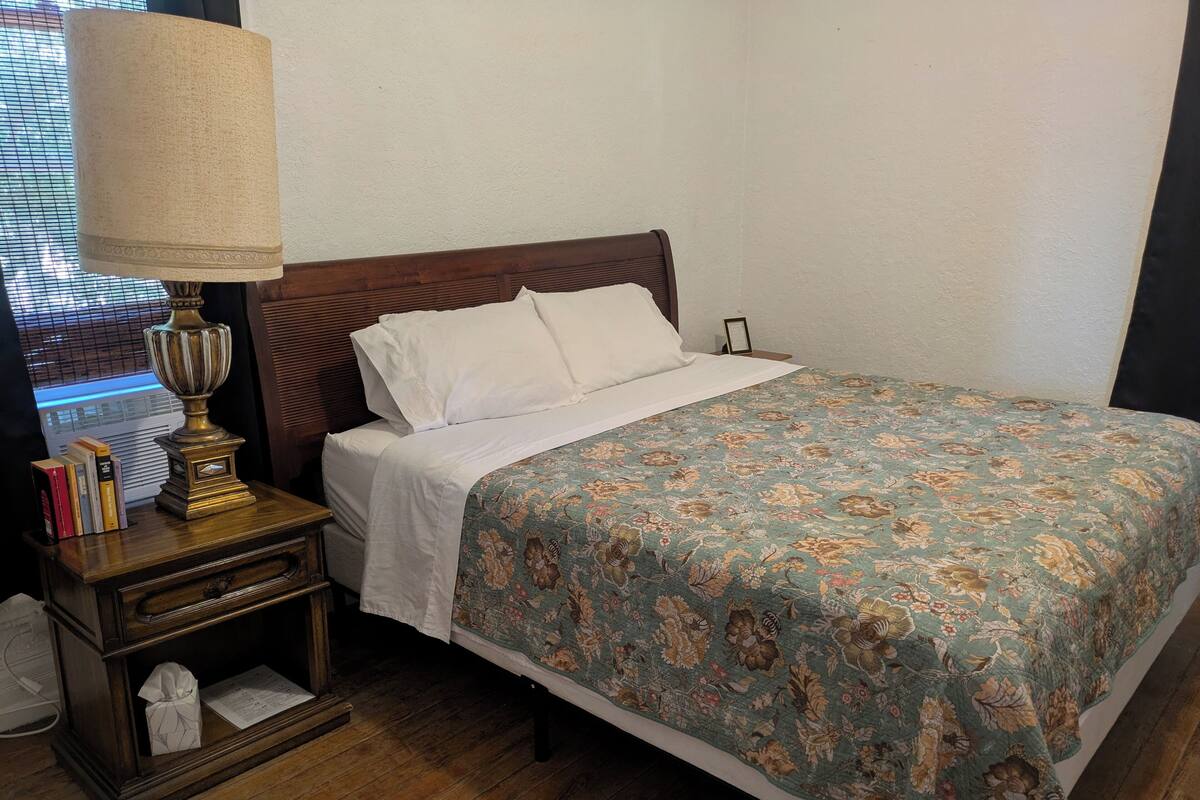 A neatly made bed with a floral bedspread, a bedside lamp, and a nightstand with books and a tissue box.