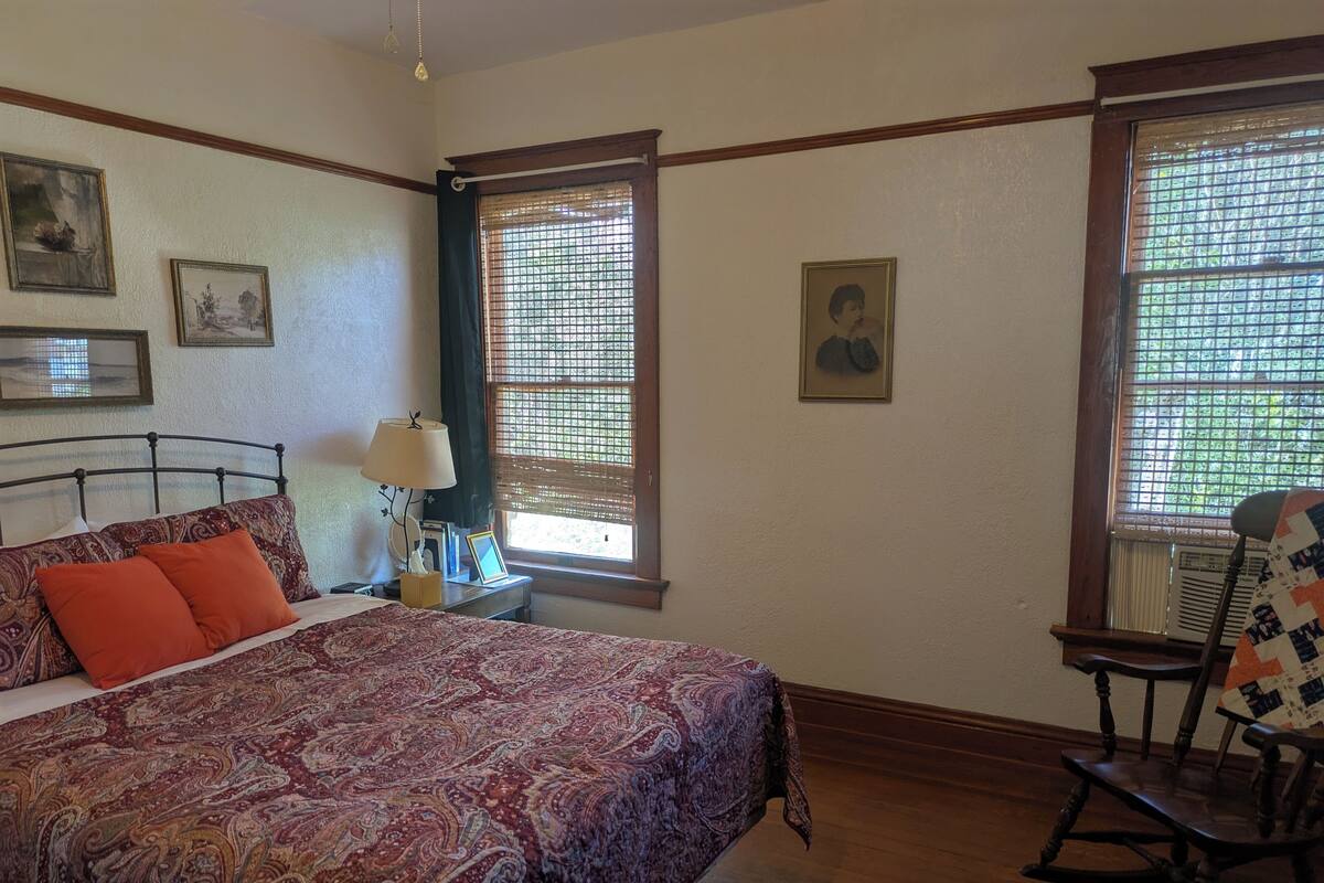 A bedroom with a large bed, a rocking chair, two windows with blinds, a bedside table with a lamp, and framed pictures on the walls.