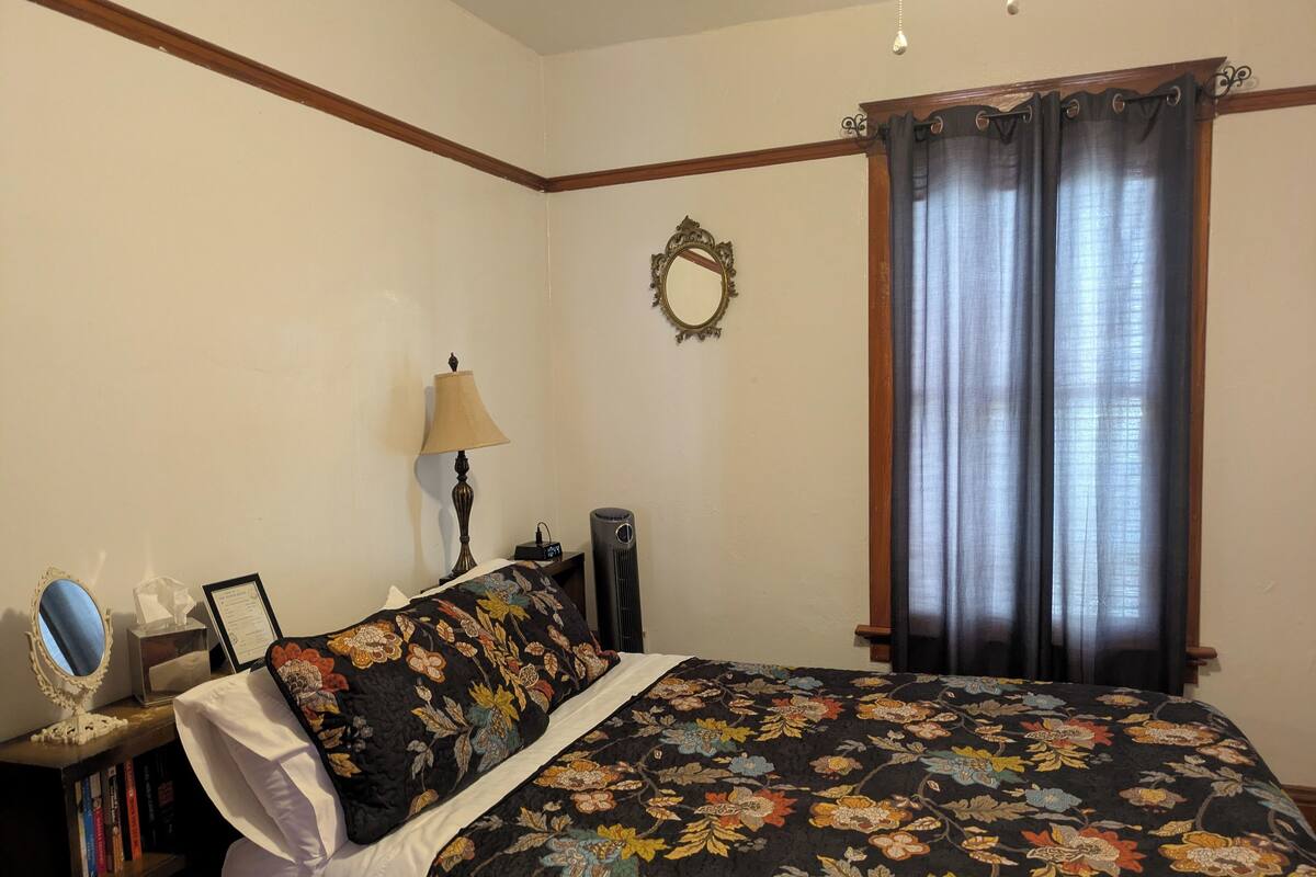A bedroom with a floral bedspread, a bedside table with a lamp, a mirror, and a framed picture.