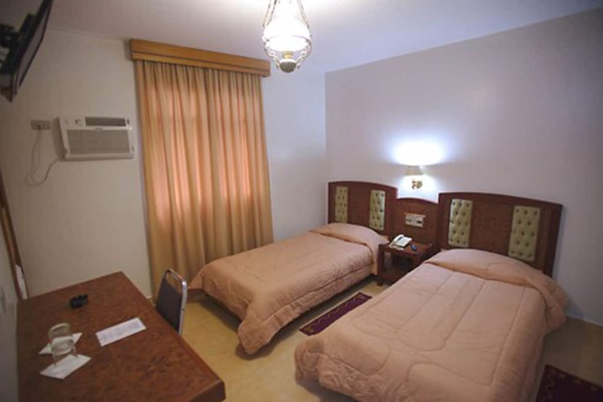 A hotel room with two single beds, a wooden desk with a telephone, and a wall-mounted air conditioning unit.