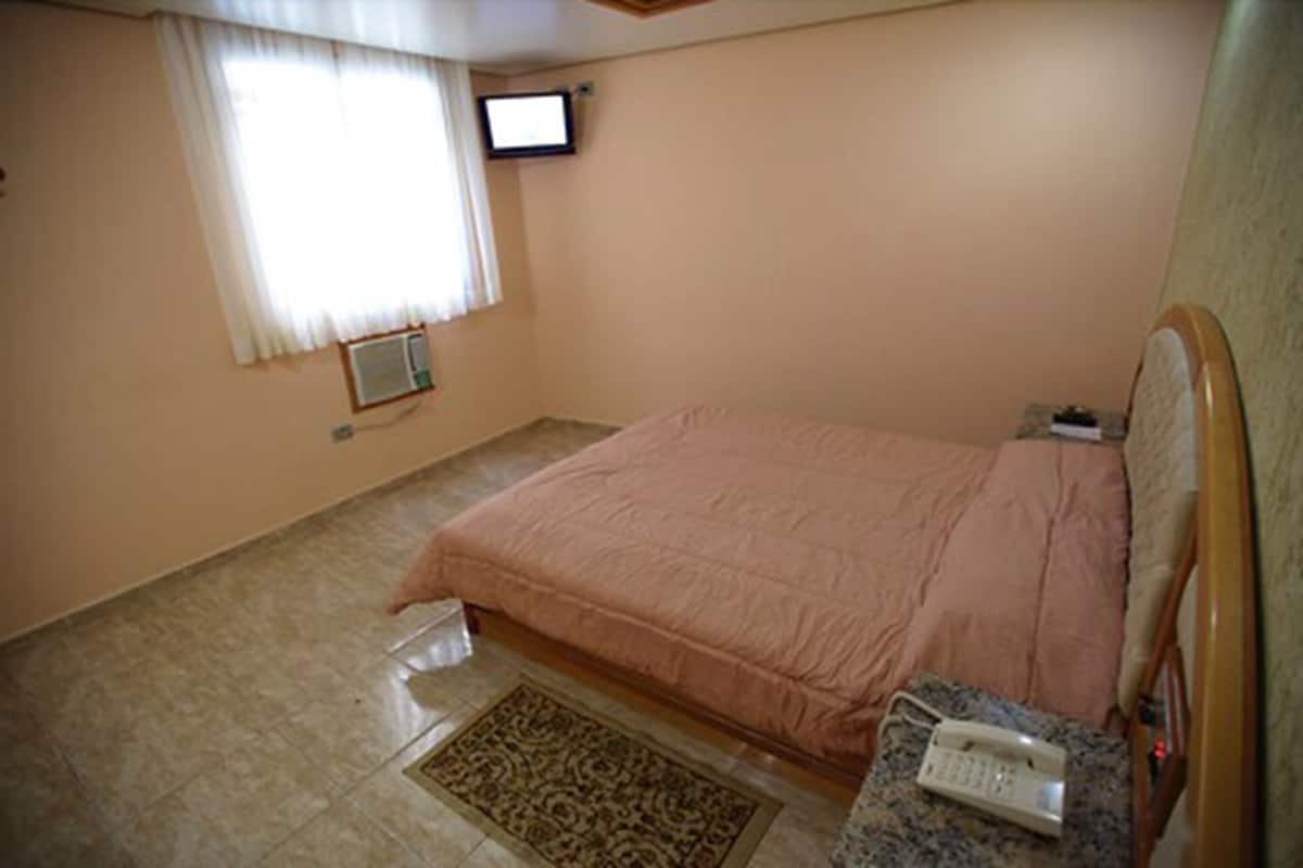 A bedroom with a bed, a television mounted on the wall, a telephone on the bedside table, and a window with curtains.