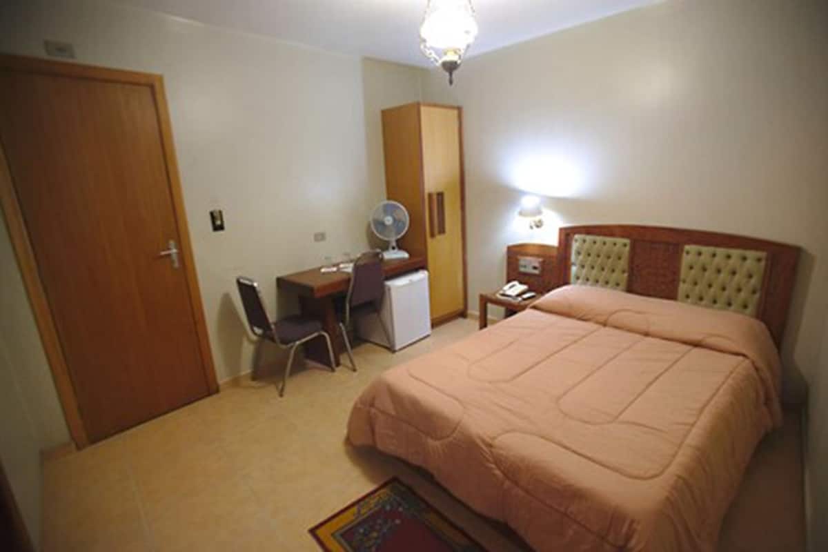 A bedroom with a bed, a desk, a chair, a wardrobe, and a nightstand with a lamp.