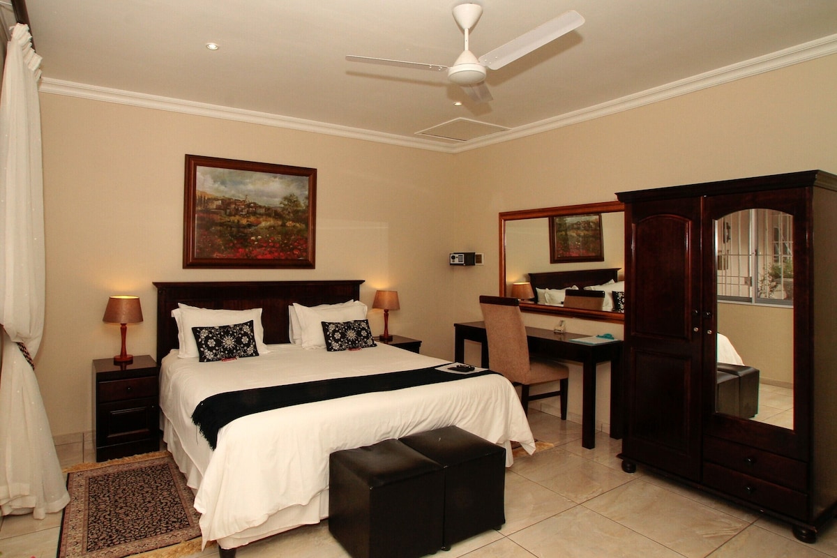 A hotel room with a bed, bedside tables, a desk, a chair, a wardrobe, a ceiling fan, and a painting on the wall.