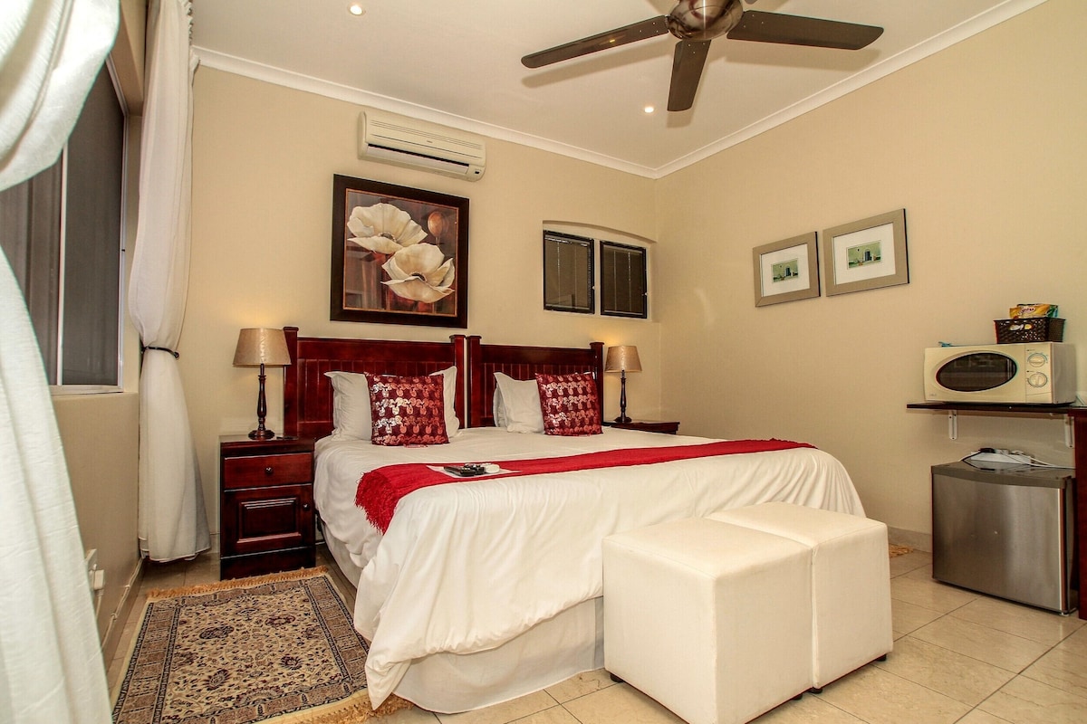 A hotel room with a bed, a nightstand, a lamp, a ceiling fan, a window, and a wall-mounted air conditioner.
