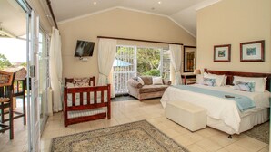 Un dormitorio con cama, una silla de madera, un sofá, televisión montada en la pared y una ventana con cortinas.