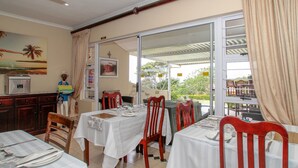 Un comedor con mesas dispuestas para dos, sillas de madera, un refrigerador y vistas al exterior a través de un ventanal.