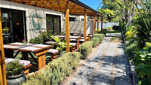 Un patio al aire libre con mesas y bancos de madera, rodeado de macetas con plantas y un sendero de piedra.