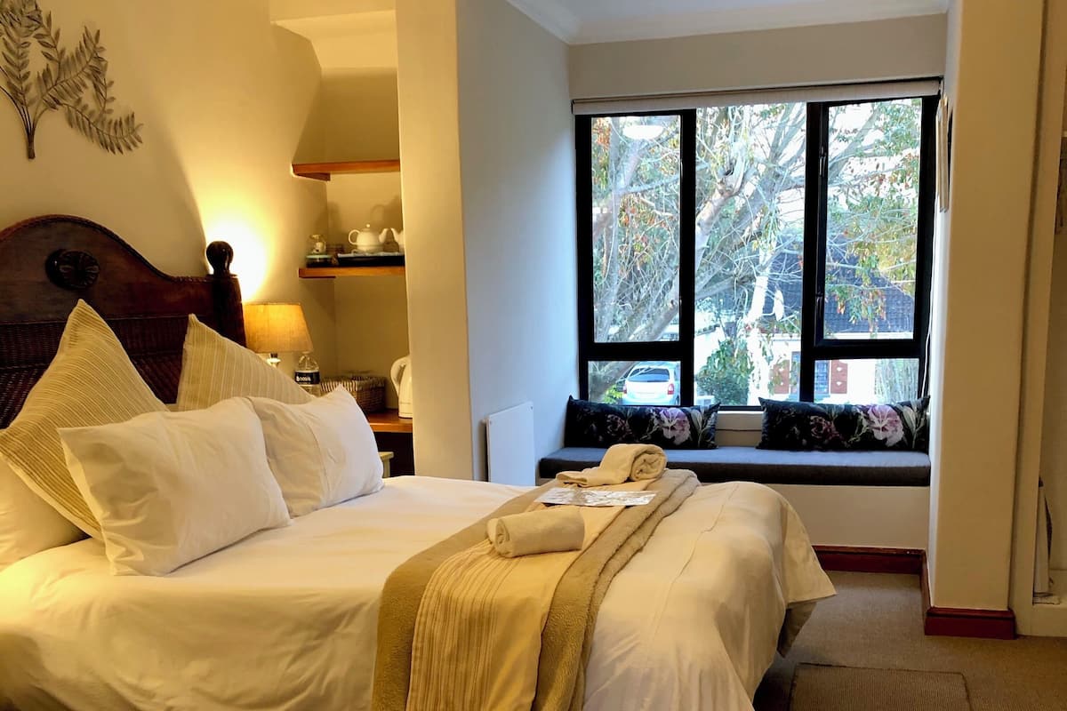 A neatly made bed with white linens and a beige throw blanket, a wooden headboard, a bedside table with a lamp, and a window with a view of trees and a car outside.