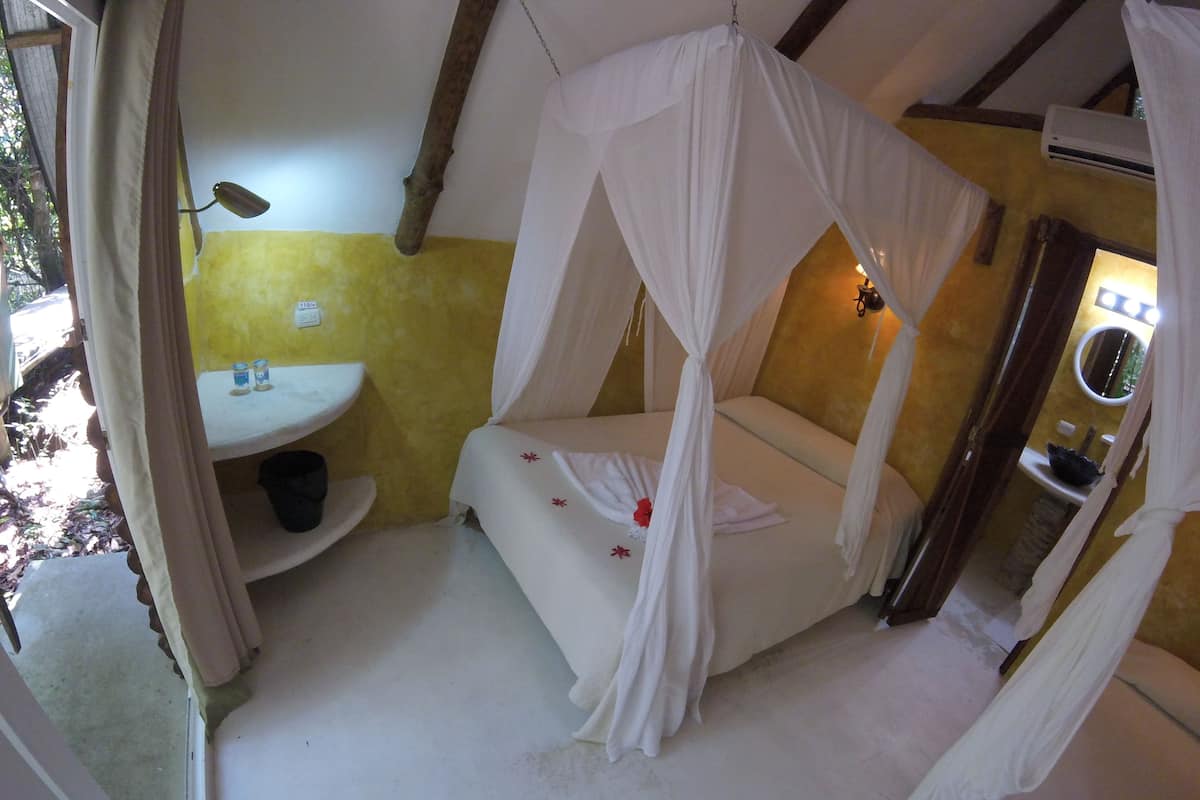 A room with a bed under a canopy, a small table with a lamp, and a sink.