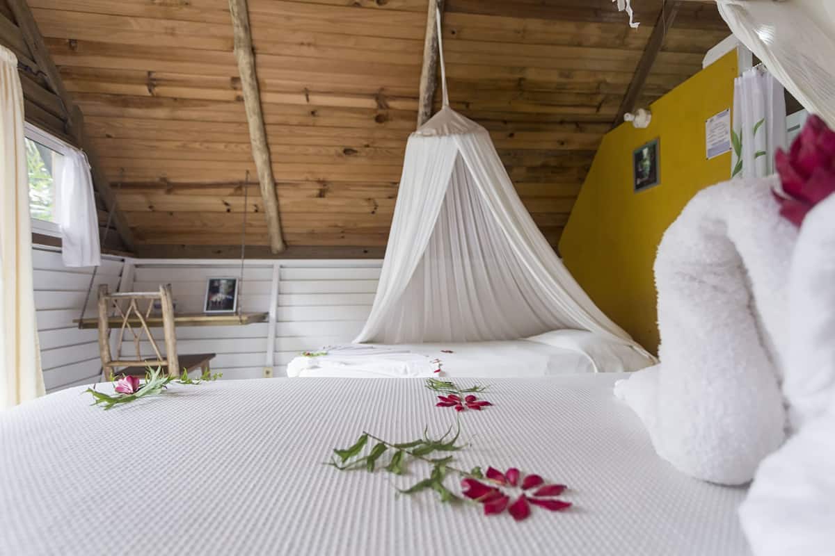 A bedroom with a bed, white bedding, a canopy, a wooden ceiling, a yellow wall, and a window.