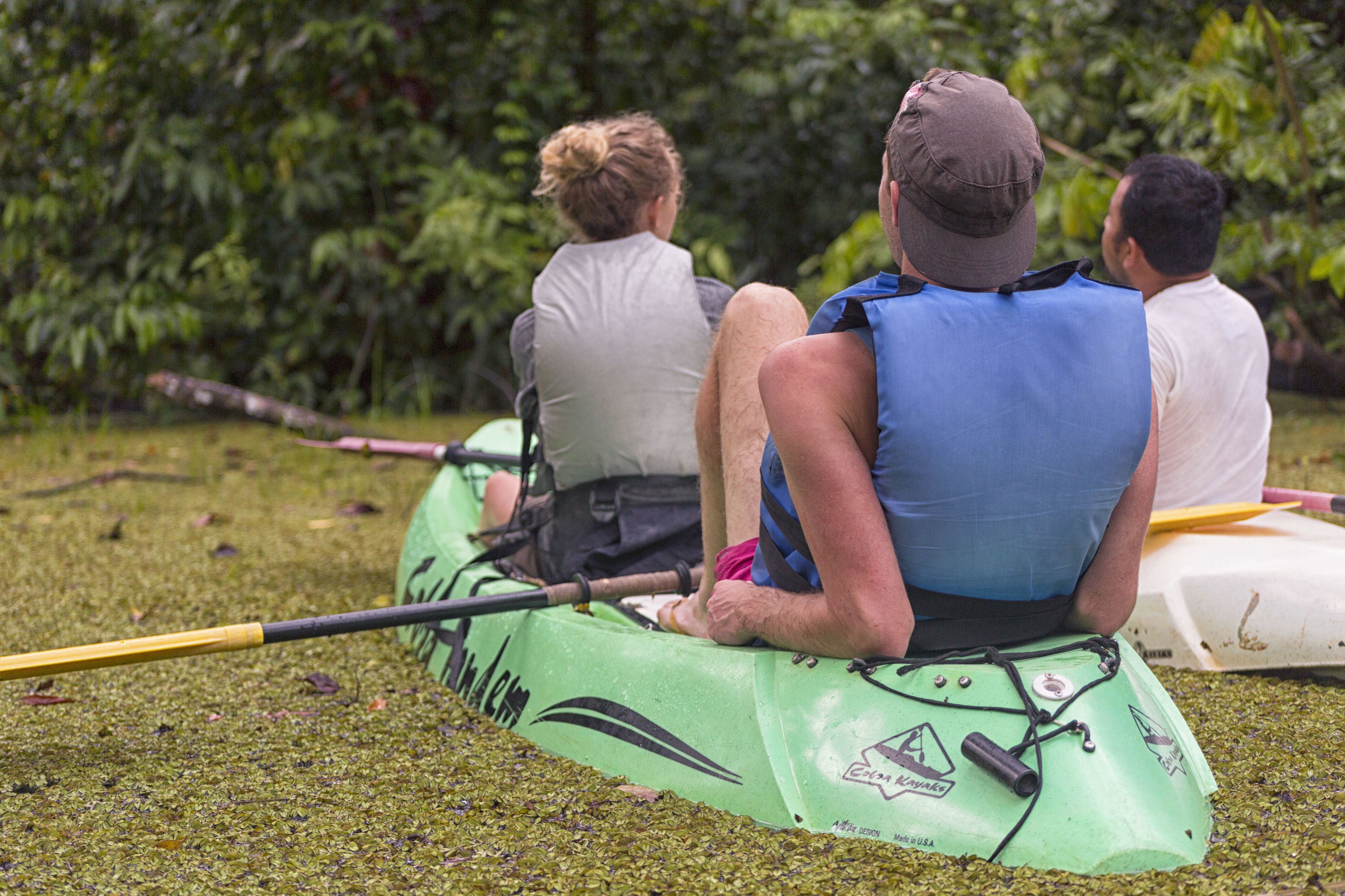 Three people sitting on a green kayak in a grassy area with dense foliage in the background.