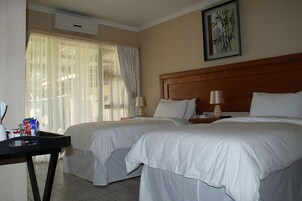 Quarto de hotel com duas camas, um painel de cama de madeira, uma mesa com abajur e uma janela com cortinas leves.