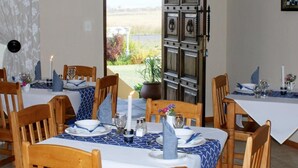 A dining area with tables set for two, featuring blue and white tablecloths, wooden chairs, and a view of a garden through an open door. - Mountain View Guest House (Glencoe)