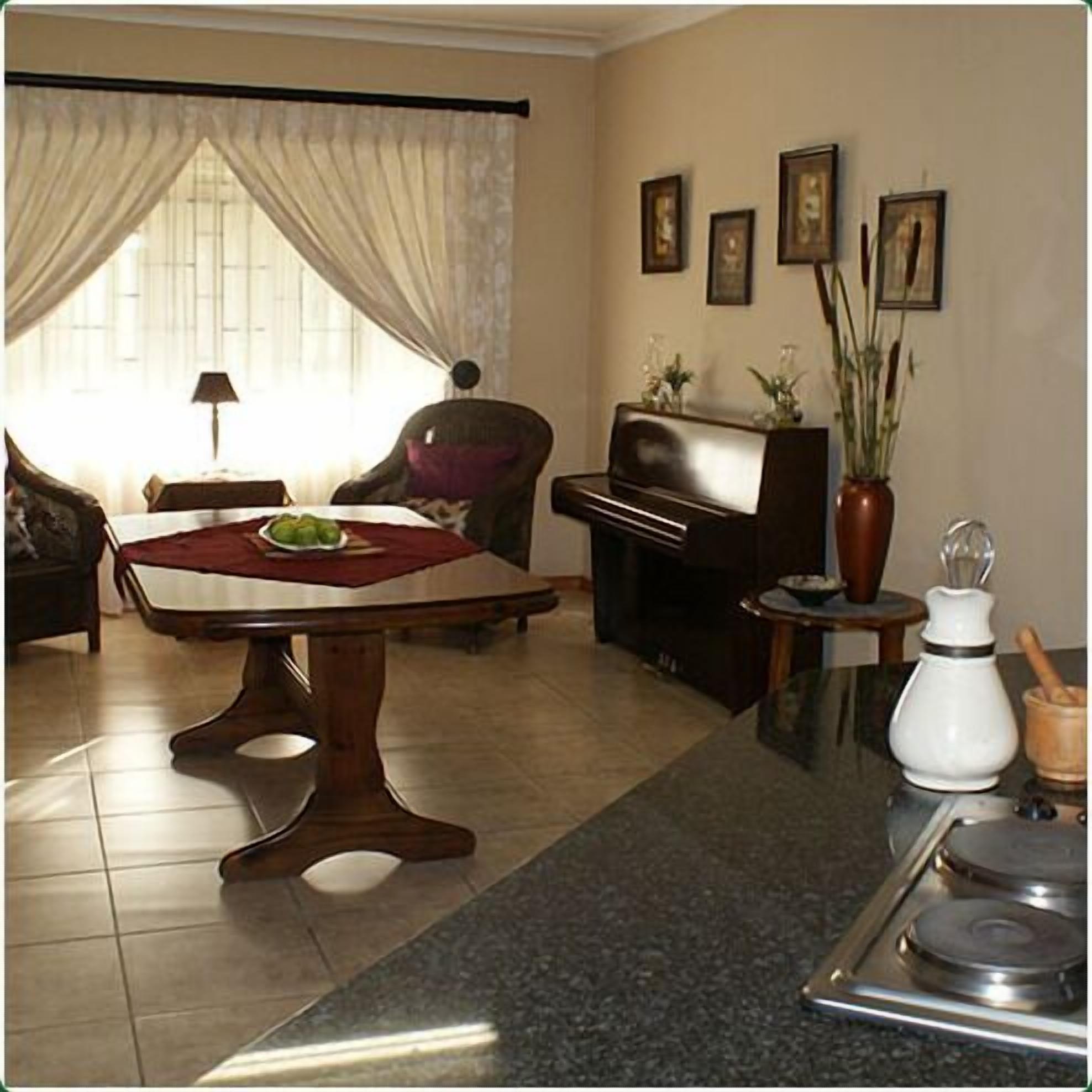 A living room with a piano, a dining table with a red placemat, a sofa, a chair, and a coffee table. There are framed pictures on the wall and a vase with plants on a side table.