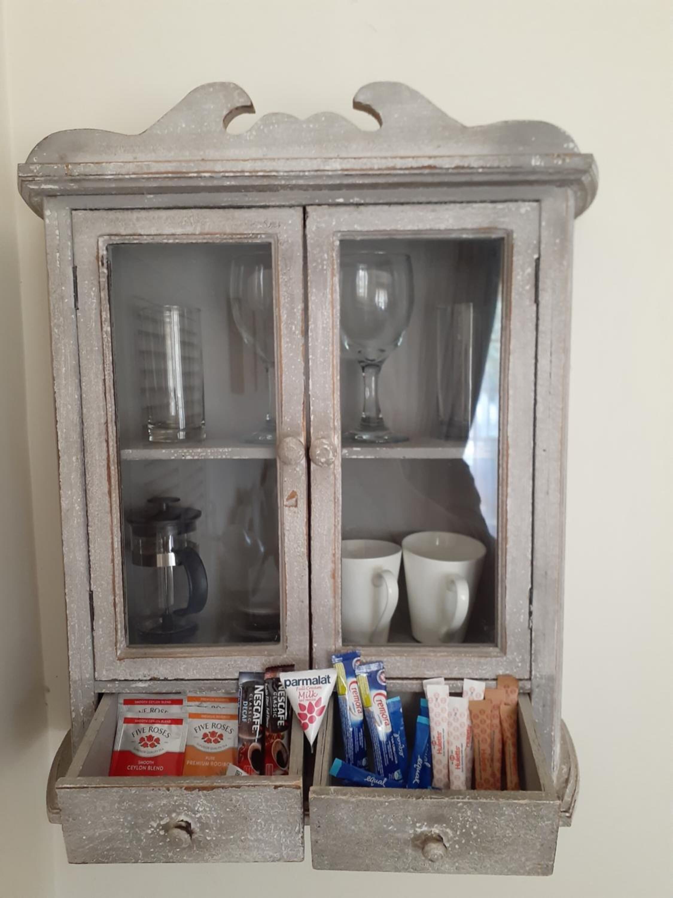 A rustic wooden cabinet with two glass doors, each containing various items like coffee makers, cups, and packaged goods.
