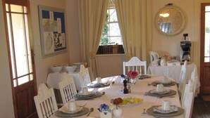 A dining room with a round table set for six, white chairs, a chandelier, and a window with curtains. - Sandstone Chameleon Guesthouse Fouriesburg (Fouriesburg)