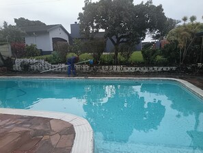A swimming pool with a stone tile border, surrounded by a stone wall and vegetation.