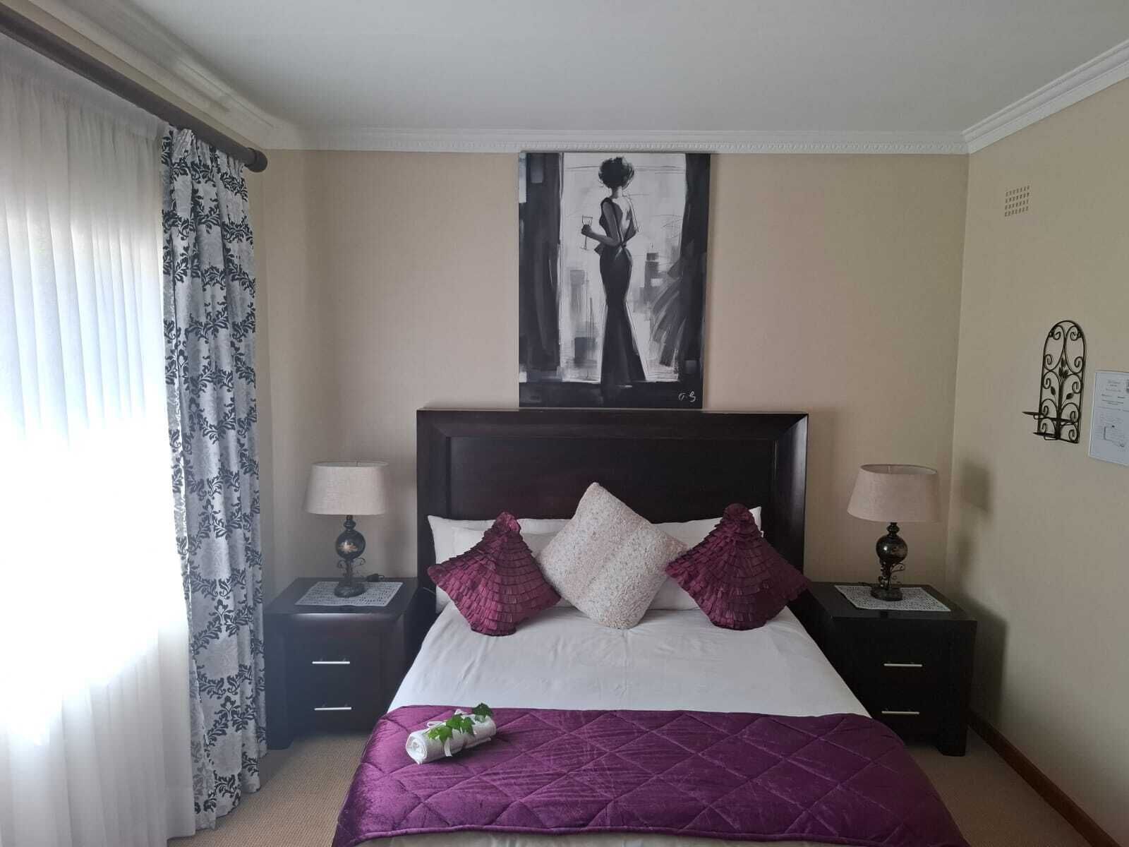 A hotel room with a bed, two bedside tables, a window with curtains, a wall-mounted artwork, and a wall-mounted light fixture.