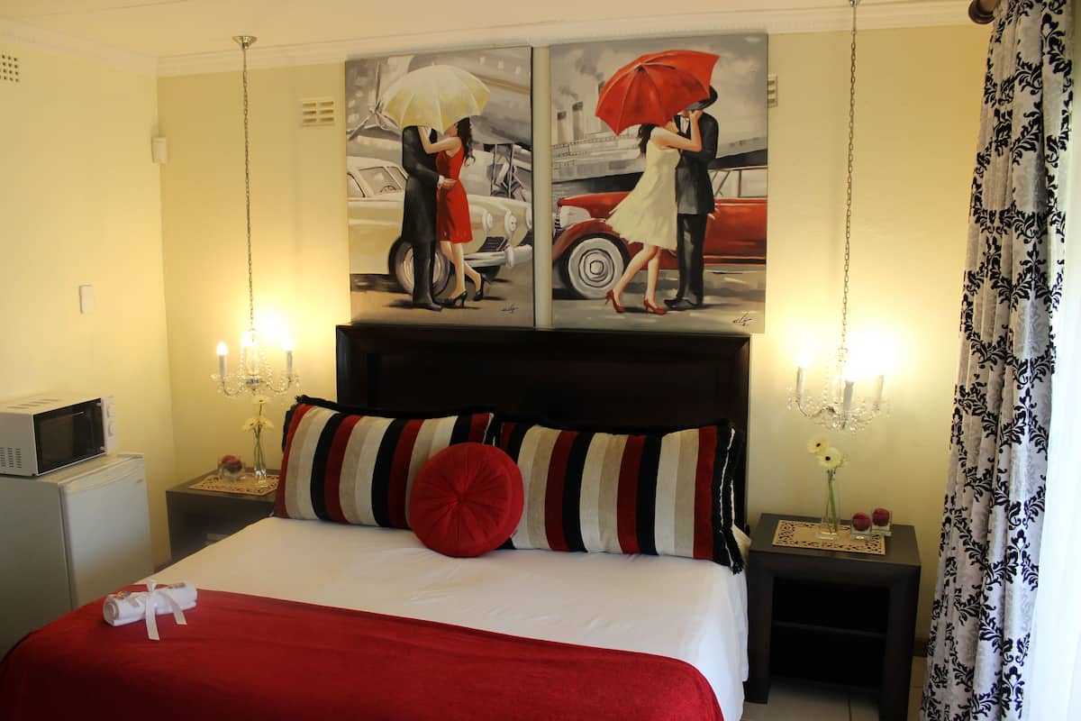 A hotel room with a large bed, a painting of a couple on a street, a microwave, a nightstand with a lamp, and a patterned curtain.
